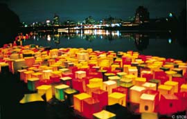 Hundreds of boxed lanters float on a river, with the city lights in the background
