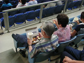 青いベンチに座り、ビールを片手に寿司を食べ、試合を楽しむ老夫婦。