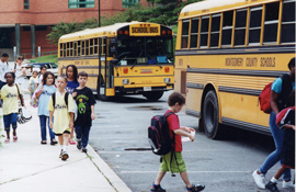 黄色いスクールバスが走る横を子供たちが歩いています。